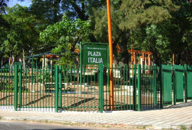 Entrada de la Plaza Italia en Asunción, con su cartel, rejas verdes y árboles